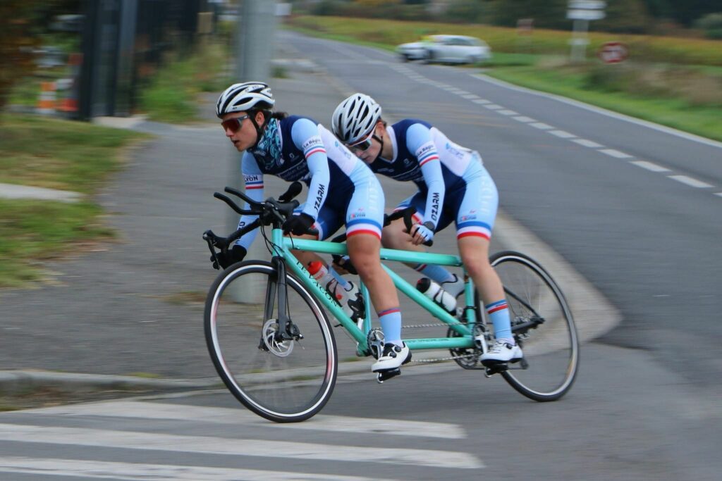 Juliette Perrochet (stoker, déficiente visuelle) et Mayliss Bonnace (pilote) souriantes, vêtues de tenues cyclistes, sur leur tandem professionnel (matériel sportif d’environ 2,50 m) en plein virage