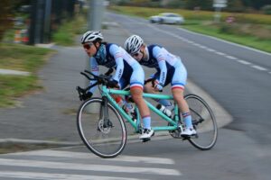 Juliette Perrochet (stoker, déficiente visuelle) et Mayliss Bonnace (pilote) souriantes, vêtues de tenues cyclistes, sur leur tandem professionnel (matériel sportif d’environ 2,50 m) en plein virage